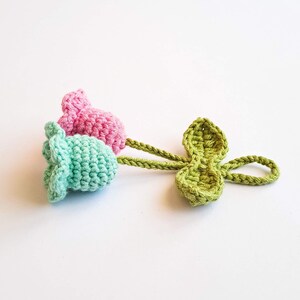 May include: Crocheted flower buds in pink and aqua blue, connected by a green stem to a green leaf. The handmade floral design is set against a white background. The crochet work is detailed and textured.