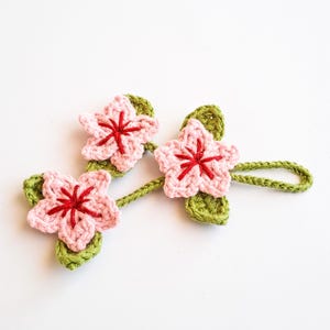May include: Crocheted floral embellishment featuring three pink flowers with red centers and green leaves. The flowers are connected by a green stem, creating a decorative element. Ideal for crafting or adding a touch of handmade charm.