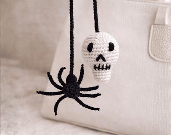 May include: Crocheted Halloween decorations featuring a black spider and a white skull. The spider has eight legs and is suspended by a black string. The skull has black eye sockets and a stitched mouth. The decorations are hanging in front of a white handbag.