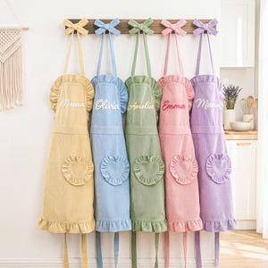 May include: Five pastel-coloured corduroy aprons: yellow, blue, green, pink, and purple. Each apron features a ruffled top, a heart-shaped pocket, and an embroidered name: Mama, Olivia, Amelia, Emma, and Mama. They hang from a wooden rack with bow details.