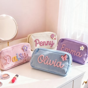Puede incluir: Bolsos de cosméticos de pana en colores pastel, cada uno con un nombre bordado en la parte delantera. Los bolsos son de color rosa, blanco, azul y morado, con adornos decorativos como flores y una mariposa. Un espejo y maquillaje en primer plano.