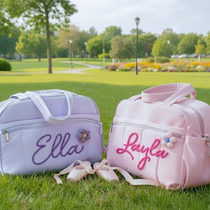 May include: Two personalized baby bags, one lavender and one pink, with the names "Ella" and "Layla" embroidered on them. Each bag has a flower accent and is paired with ballet slippers. The bags are on green grass.
