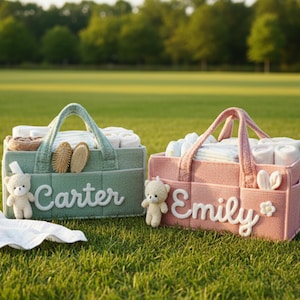 May include: Two baby gift baskets on green grass. One is mint green with the name "Carter" and a teddy bear. The other is pink with the name "Emily" and a teddy bear. Both baskets contain baby supplies.