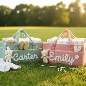May include: Two personalized baby caddies, one green and one pink, sit on grass. The green caddy, labeled "Carter," holds baby supplies. The pink caddy, labeled "Emily," also holds baby supplies. Each caddy is 15 inches long, 10 inches wide, and 7 inches tall.
