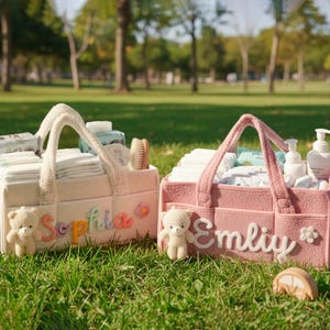 May include: Two personalized baby diaper caddies in a park setting. One is cream-colored with the name "Sophia" and the other is pink with the name "Emily". Each caddy contains baby supplies and a small teddy bear.