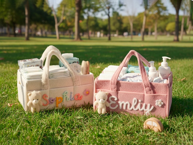 May include: Two personalized baby diaper caddies in a park setting. One is cream-colored with the name "Sophia" and the other is pink with the name "Emily". Each caddy contains baby supplies and a small teddy bear.