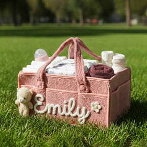 May include: A pink baby diaper bag with the name "Emily" in white script. The bag contains diapers, a bottle, socks, and a small teddy bear. The bag is sitting on green grass.