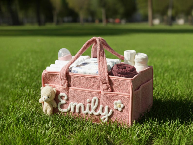 May include: A pink baby diaper bag with the name "Emily" in white script. The bag contains diapers, a bottle, socks, and a small teddy bear. The bag is sitting on green grass.