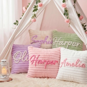 May include: A collection of five square, ribbed, plush throw pillows in pastel shades of lavender, light pink, sage green, and white. Each pillow features a soft, raised, cursive name embroidered on the front, including the names Sophie, Harper, Olivia, and Amelia.