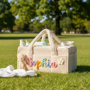 May include: Cream-colored baby bag with a teddy bear and the name "Sophia" in colorful letters. The bag has a fluffy texture and is filled with baby essentials. A white baby blanket rests on the grass.