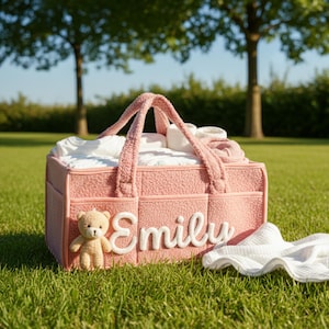 May include: A pink baby diaper caddy with the name "Emily" in white script. The caddy has a teddy bear attached and is filled with baby essentials. A white cloth is next to the caddy on the grass. The caddy has a handle for easy carrying.