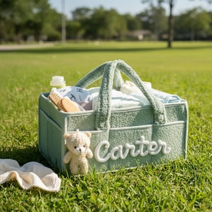 May include: A light green baby basket with the name "Carter" in white script. The basket is filled with baby items, including a bottle, brush, and soft toys. A small teddy bear sits next to the basket on the grass.