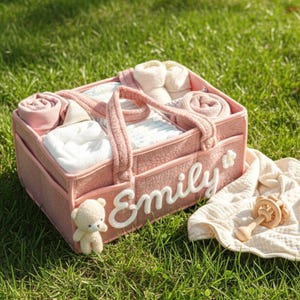 May include: A pink baby gift basket filled with diapers, rolled blankets, socks, and other baby essentials. The basket has the name "Emily" in white script. A small teddy bear and a wooden rattle are nearby.