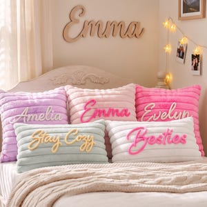 Peut inclure: Cinq coussins décoratifs en peluche côtelée aux couleurs pastel : lavande, rose clair, rose vif, vert menthe et blanc. Les coussins présentent des inscriptions en relief en lettres cursives : Amelia, Emma, Evelyn, Stay Cozy et Besties.