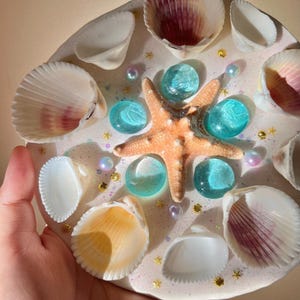 May include: A decorative, round resin piece featuring a starfish, seashells, and turquoise glass stones. The piece is adorned with gold star-shaped glitter and iridescent pearls, creating a coastal theme. The seashells have white and purple hues.