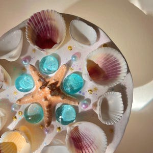 May include: A round, white decorative piece adorned with seashells, a starfish, and turquoise glass beads. The piece is embellished with small gold star-shaped accents and iridescent pearls, creating a coastal theme.