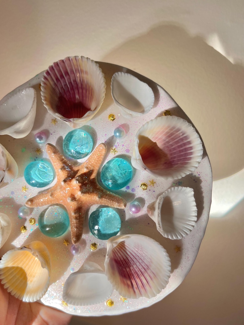 May include: A round, white decorative piece adorned with seashells, a starfish, and turquoise glass beads. The piece is embellished with small gold star-shaped accents and iridescent pearls, creating a coastal theme.