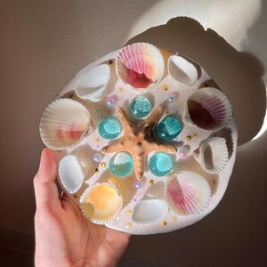 May include: A round, decorative piece featuring a starfish, seashells, and blue glass beads. The seashells are white, pink, and yellow. The piece is set on a white, glittery base. A unique, beach-themed decorative item.