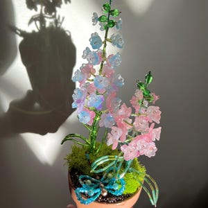 Pode incluir: Um arranjo floral de contas artesanal num vaso de terracota. As flores são rosa, azuis e transparentes, com caules e folhas verdes. Uma libélula de contas está sentada no musgo. O vaso tem cerca de 10 cm de diâmetro.