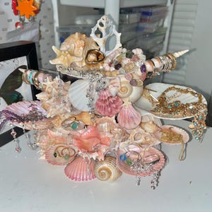 K&ouml;nnte beinhalten: Ein einzigartiger Muschelschmuckhalter mit einer Vielzahl von Muscheln in Rosa-, Creme- und Wei&szlig;t&ouml;nen. Der Halter ist mit Ringen, Halsketten und anderem Schmuck verziert und schafft eine dekorative und funktionale Pr&auml;sentation. Der Halter steht auf einer wei&szlig;en Oberfl&auml;che.