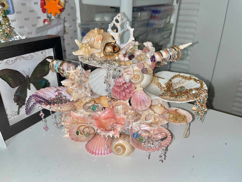 K&ouml;nnte beinhalten: Ein einzigartiger Muschelschmuckhalter mit einer Vielzahl von Muscheln in Rosa-, Creme- und Wei&szlig;t&ouml;nen. Der Halter ist mit Ringen, Halsketten und anderem Schmuck verziert und schafft eine dekorative und funktionale Pr&auml;sentation. Der Halter steht auf einer wei&szlig;en Oberfl&auml;che.