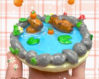 May include: A miniature clay scene of two brown capybaras floating in a blue pond with green lily pads. The pond is surrounded by gray rocks and green plants. There are orange fruit on top of the capybaras and in the pond.