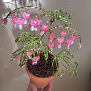 May include: A handcrafted beaded plant in a small terracotta pot. The plant features green beaded leaves and stems, with pink heart-shaped flowers. The plant is held in a person's hand, with a neutral background.