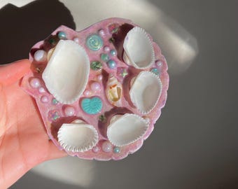 May include: A heart-shaped, pink resin piece adorned with white seashells, pearls, and small decorative elements. The resin has a glittery finish, and the seashells are arranged on the surface. The piece is held in a hand.