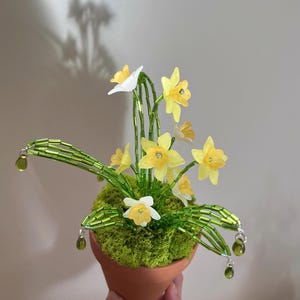 May include: A handcrafted floral arrangement featuring yellow and white daffodils in a small terracotta pot. The arrangement includes green beaded stems and leaves, with green teardrop-shaped accents. The pot is filled with green moss.