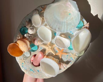 May include: A decorative, round seashell and resin art piece. The piece is adorned with various seashells, starfish, and turquoise glass beads. The resin base is a light beige color with glitter accents. The piece is held by a hand.
