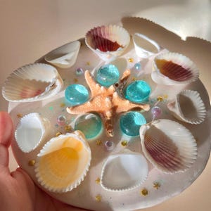 May include: A round, decorative piece featuring a starfish, seashells, and blue glass beads. The seashells are white, with some having red and yellow accents. Gold star-shaped glitter and iridescent glitter are scattered around the design.