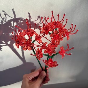 May include: A handcrafted red flower arrangement with green stems and clear accents. The flower is held in a hand against a white background, casting a shadow. The arrangement is made of small, detailed components.