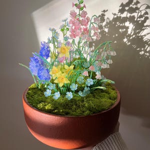 May include: A terracotta pot filled with green moss and a variety of glass flowers in shades of blue, pink, yellow, and green. The flowers are arranged in a bouquet-like shape, with the pink flowers at the top and the blue flowers at the bottom.