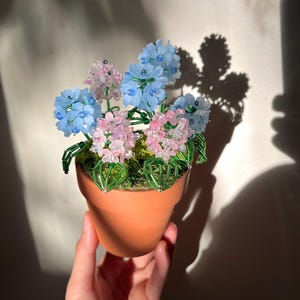 May include: A small terracotta pot filled with beaded flowers in shades of blue and pink. The flowers have green beaded stems and sit on green moss. The pot is held in a hand, with sunlight casting shadows.