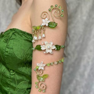 Peut inclure: Un bracelet de bras en fil doré orné de feuilles en verre vert, de petites fleurs blanches et de perles vert clair. Le design présente des spirales en fil métallique, des clochettes blanches pendantes et une fleur centrale avec une perle.