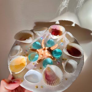 May include: A round, clear resin coaster with embedded seashells, blue glass beads, a starfish, and gold glitter accents. The coaster is held in a hand and has a coastal theme.