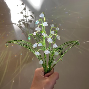 May include: A handmade bouquet of lily of the valley flowers. The stems and leaves are made of green wire, and the delicate bell-shaped flowers are white. The bouquet is held in a hand, with a soft background.