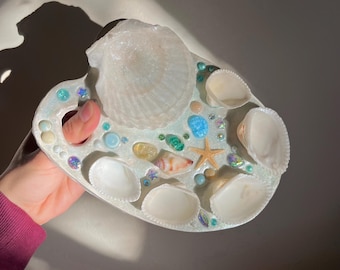 May include: A decorative, seashell-themed tray. The tray is white with a light blue border and is adorned with various seashells, small beads, and a starfish. The tray is held by a person.
