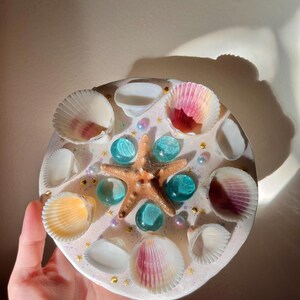 May include: A round, decorative piece featuring a starfish, seashells, and blue glass beads. The seashells are white, pink, and yellow, and are arranged around the edges. Gold star-shaped accents and small pearls are also included.