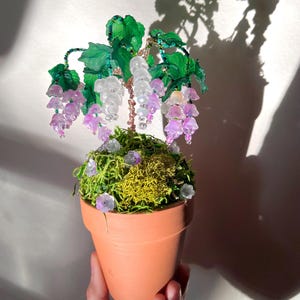May include: A beaded flower arrangement in a terracotta pot. The flowers are pink and white and the leaves are green. The arrangement is sitting on a table.