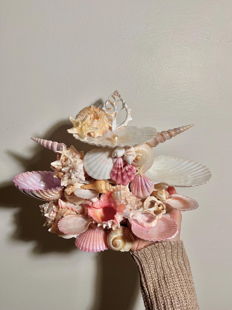 May include: A decorative arrangement of seashells in a variety of colours, including pink, white, and tan. The display features conch shells, scallop shells, and other types, creating a layered, textured look. The shells are held in a hand.