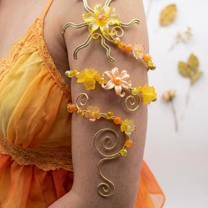 May include: A gold-tone wire arm cuff featuring a sun-shaped centrepiece with yellow petals, accented by smaller orange and yellow floral beads and spiralling wire details that wrap around the upper arm in a decorative, multi-layered design.