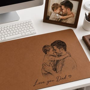 May include: A brown desk mat featuring a sketch of a father and child embracing, with the words "Love you Dad." A framed photograph of a father and child is on the desk. A mug with "Dad" is on the desk.