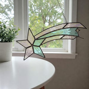 May include: A stained glass window hanging with a shooting star design. The star is a five-pointed star with a green and white iridescent finish. The tail of the shooting star is a long, curved shape with a green and white iridescent finish.