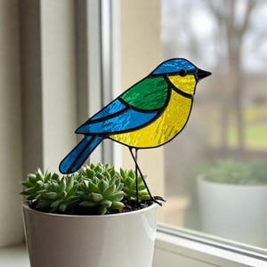 Op de afbeelding: Een blauwe glas-in-loodvogel met groene en gele accenten, die op een metalen standaard staat. De vogel staat voor een potplant met vetplanten.
