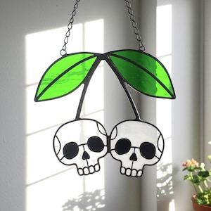 May include: Stained glass cherry design with two skull faces, black outlines, and green leaves. The decorative piece hangs from a chain. The skulls have black eye sockets and a row of teeth. The leaves are a bright green color.