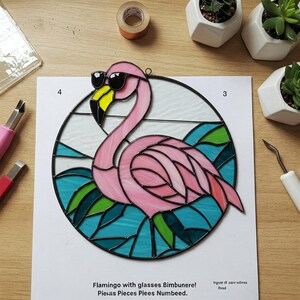 Puede incluir: Un flamenco de vidrio coloreado con gafas de sol, de pie en una escena de agua azul y verde. El flamenco es rosa con contornos negros y las gafas de sol son negras. La pieza es circular y tiene un marco negro.
