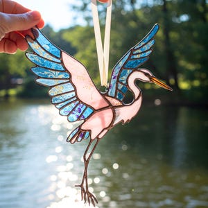 May include: A stained glass ornament of a pink and blue great egret with its wings spread. The ornament is hanging from a white ribbon.