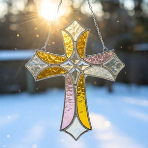 May include: A stained glass cross with a silver chain for hanging. The cross features a variety of colors, including gold, pink, and clear glass. The sun shines through the cross, creating a beautiful display of light and color. A decorative religious item.