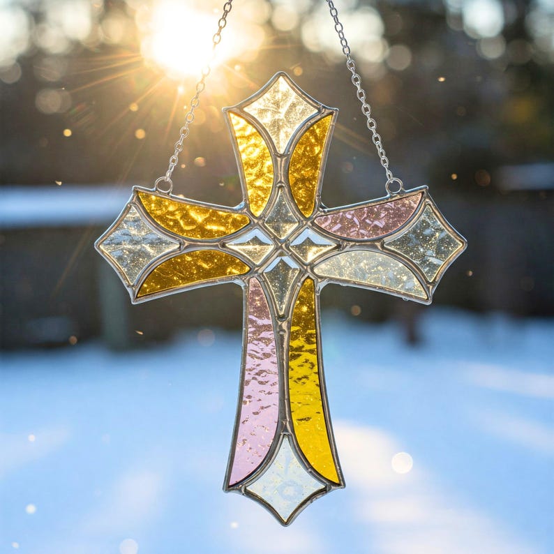 K&ouml;nnte beinhalten: Ein Buntglaskreuz mit einer silbernen Kette zum Aufh&auml;ngen. Das Kreuz hat eine Vielzahl von Farben, darunter Gold, Rosa und klares Glas. Die Sonne scheint durch das Kreuz und erzeugt ein sch&ouml;nes Spiel von Licht und Farbe. Ein dekorativer religi&ouml;ser Artikel.